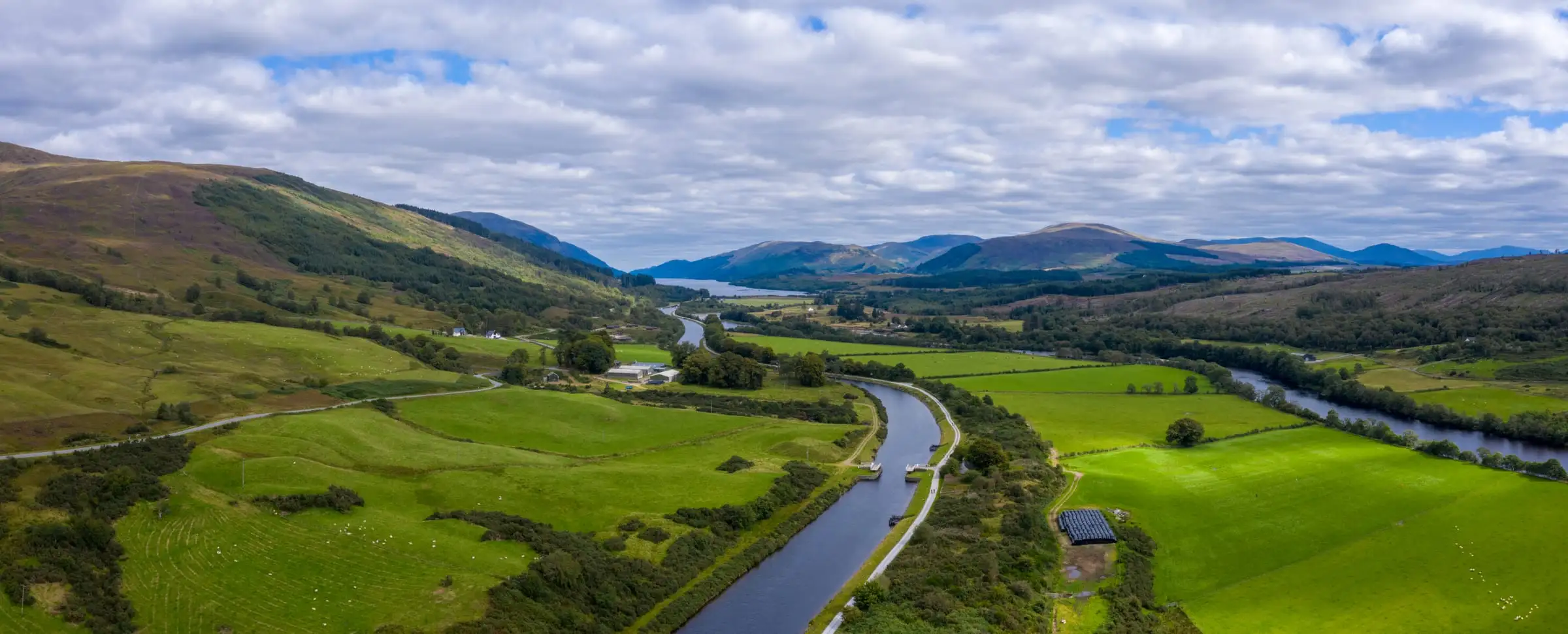 A canal running straight through a green Highland valley beside a winding river, with fields, woodland and mountains under a cloudy sky.