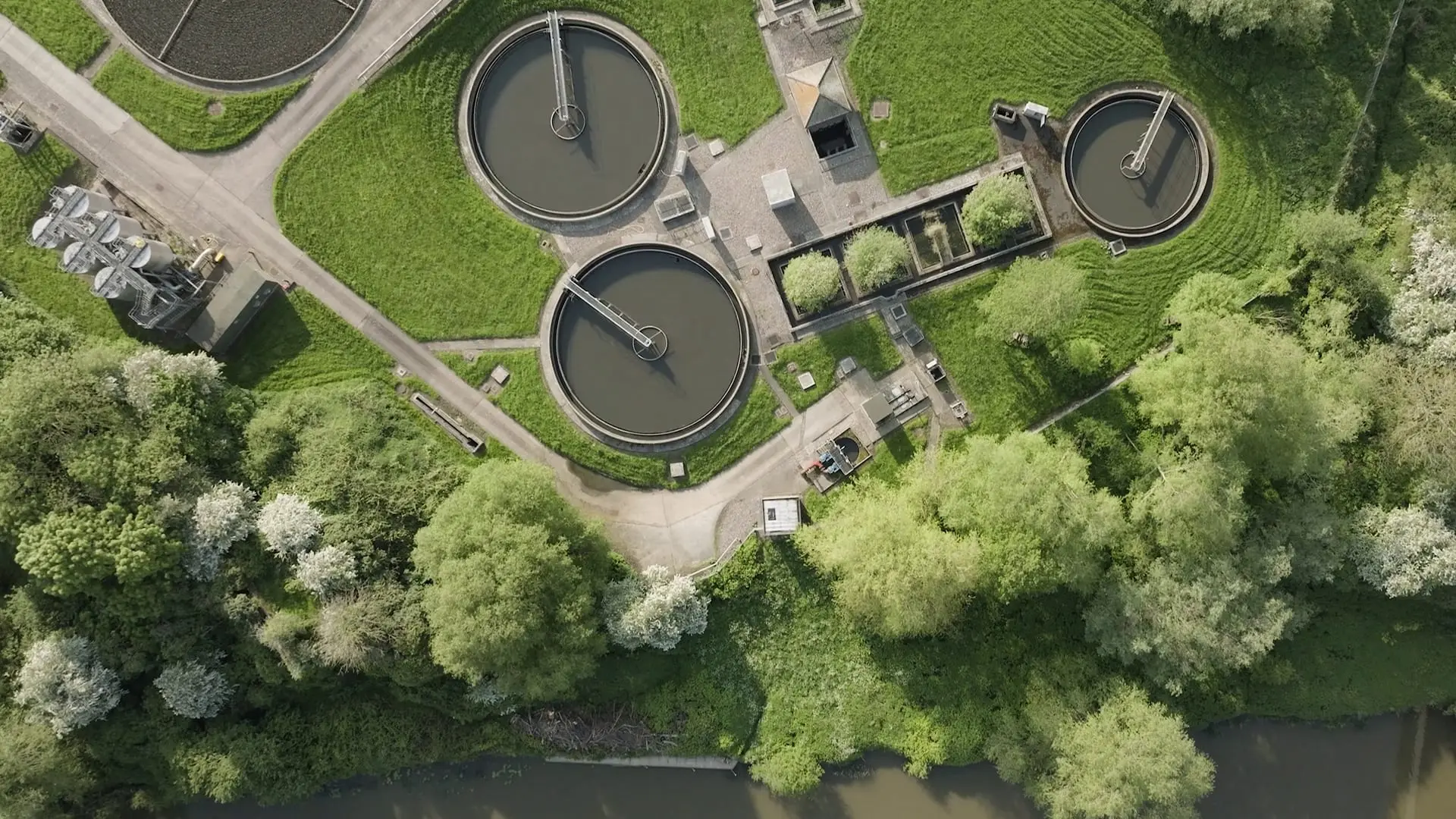 A water treatment works seen from directly above: circular settlement tanks set in mown grass, with a belt of trees between the works and a river.