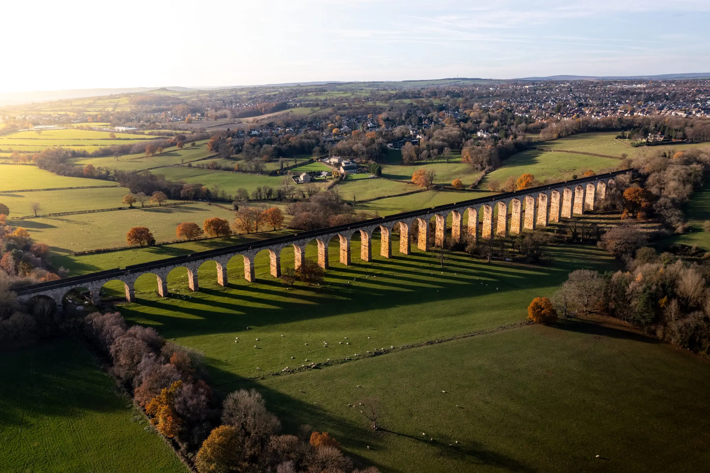 A long stone railway viaduct crossing green farmland in low sun, its arches casting shadows across the fields, with a town and hills beyond.