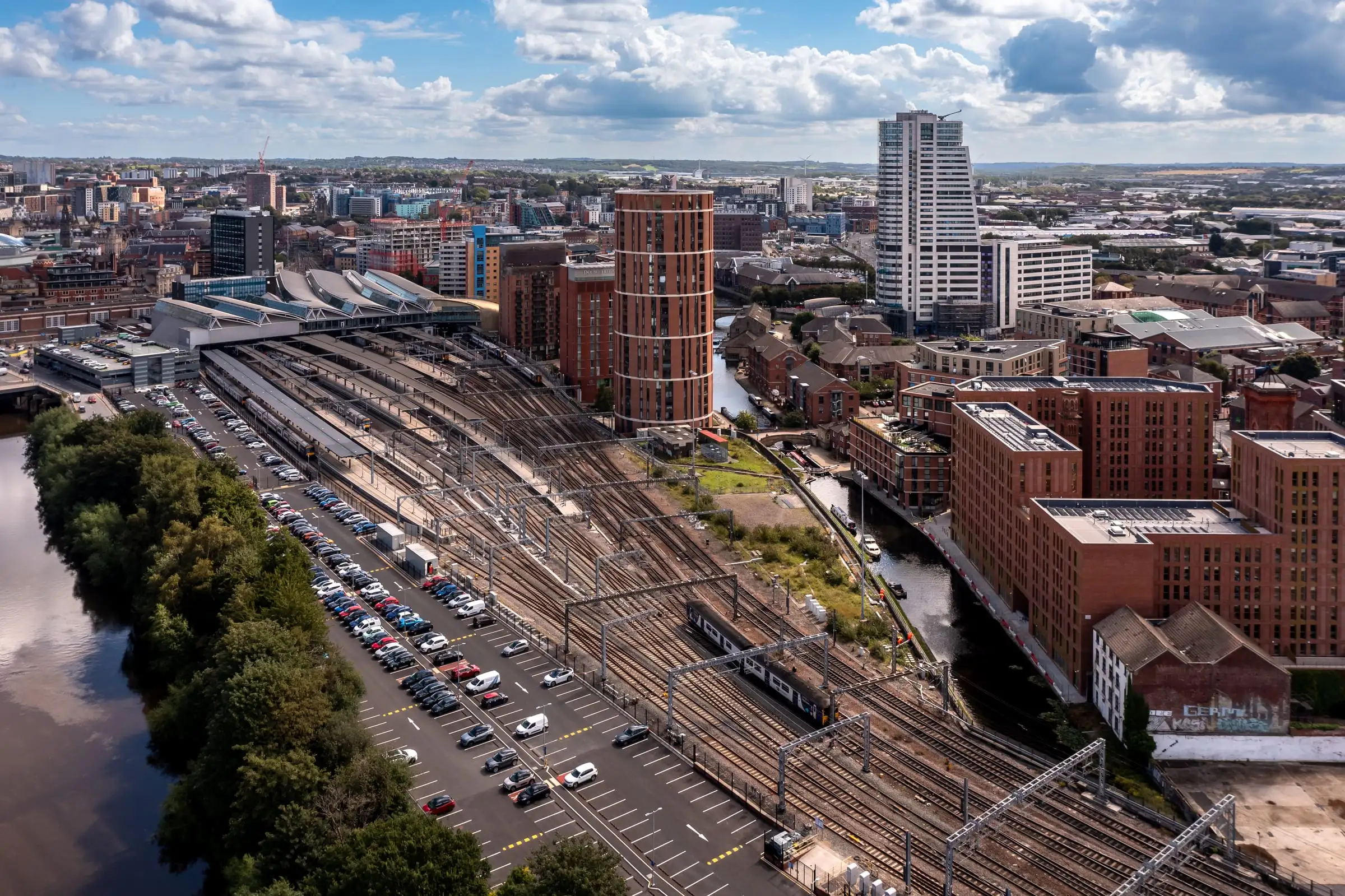 A city railway station from the air: platforms and tracks fanning out beside a river and a car park, with brick apartment blocks and towers beyond.